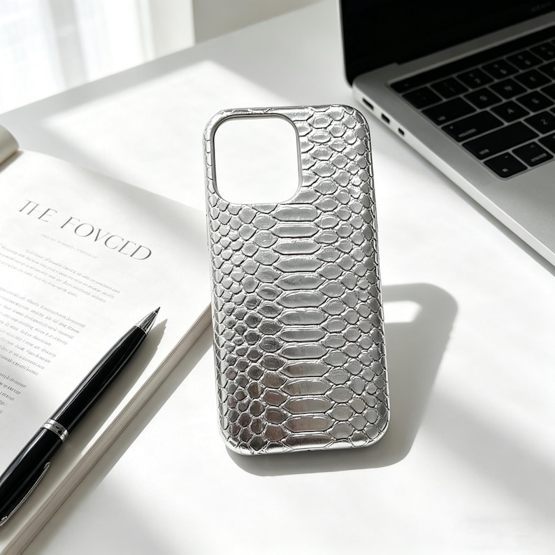 May include: Silver phone case with a snakeskin texture. The case has a cutout for the camera and buttons. The phone case is on a white surface next to a laptop and an open book with the text "THE LOVCED".