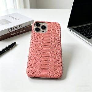 May include: A coral-colored phone case with a snakeskin texture. The case has a camera cutout and is next to a laptop, a pen, and a book titled "Craft". The case is designed to protect a smartphone.