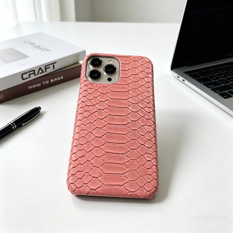 May include: A coral-colored phone case with a snakeskin texture. The case has a camera cutout and is next to a laptop, a pen, and a book titled "Craft". The case is designed to protect a smartphone.