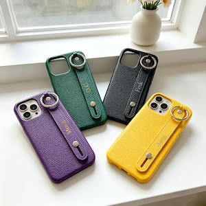 May include: Four colorful phone cases: purple, green, black, and yellow. Each case has a textured finish, a strap with a metal ring, and a small button. The straps have gold lettering: "Mom", "D.C.", "Fmk", and "Aneg".