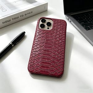 May include: A burgundy phone case with a textured, snake-skin pattern. The case is on a white surface next to a black pen, a book with the title "HOW TOW TO BAKE", and a laptop.