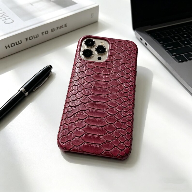 May include: A burgundy phone case with a textured, snake-skin pattern. The case is on a white surface next to a black pen, a book with the title "HOW TOW TO BAKE", and a laptop.