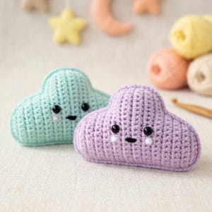 May include: Two handmade crochet cloud-shaped toys. One is light blue, the other is lavender. Both have black eyes and stitched smiles. The clouds are on a neutral background, with yarn and other crochet elements in the background.