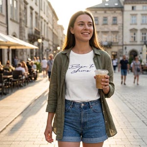 May include: A woman wearing a white t-shirt with the text "Born to Shine" in navy blue script, paired with denim shorts and an olive green button-down shirt. She is holding a clear plastic cup with a brown beverage.
