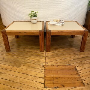 May include: Two wooden end tables with light beige tops. The tables are side by side on a wood floor.