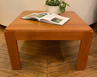 May include: A square, wooden coffee table with rounded edges and four legs. The table is made of light brown wood and has a natural finish. A book and a potted plant are on top of the table.