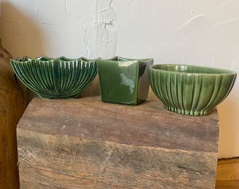 May include: Three green ceramic planters of varying shapes sit on a weathered wooden surface. The planters include a scalloped bowl, a square pot, and an oval bowl, all in a glossy, dark green glaze. The background is a textured white wall.