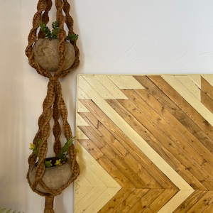 May include: A brown macrame plant hanger with two stone-colored pots, each holding greenery and small yellow flowers. The background features a wooden panel with a chevron pattern in shades of brown and cream.