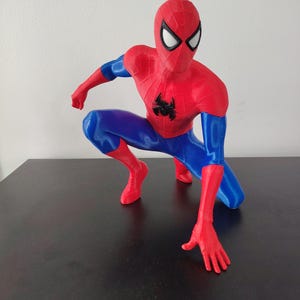 May include: A red and blue Spider-Man figurine in a crouching pose. The figure has a red mask with white eye lenses, a black spider emblem on the chest, and blue accents on the arms and legs. The figurine is on a dark surface.