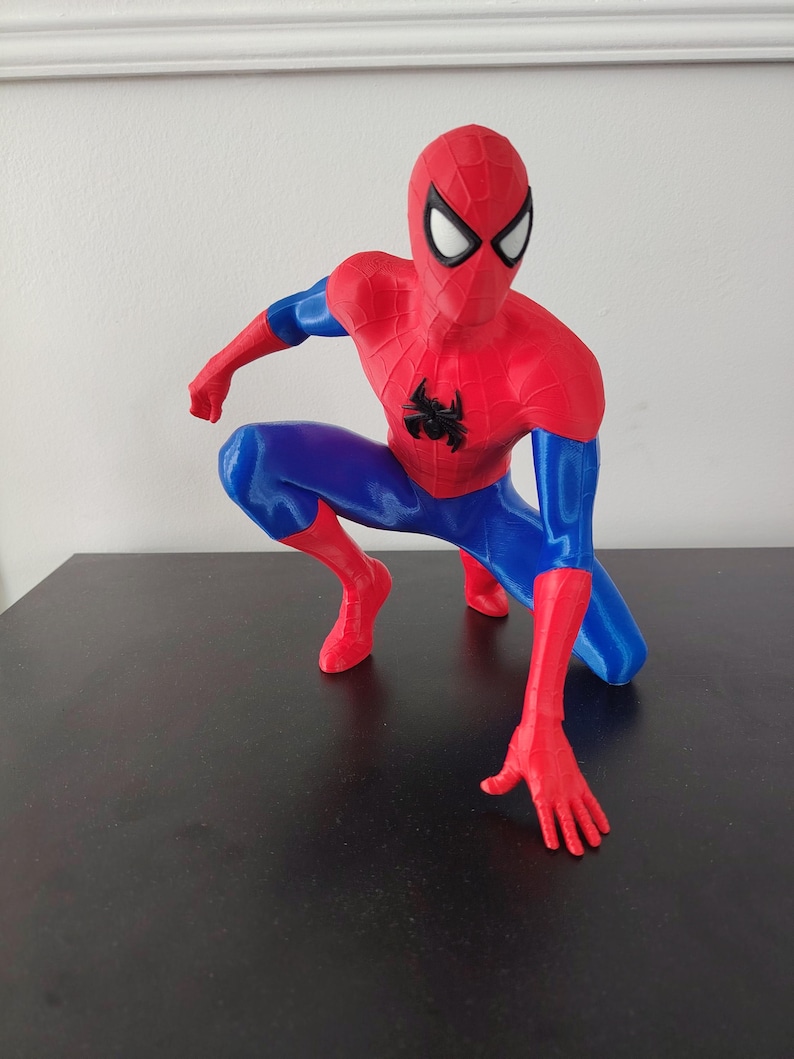 May include: A red and blue Spider-Man figurine in a crouching pose. The figure has a red mask with white eye lenses, a black spider emblem on the chest, and blue accents on the arms and legs. The figurine is on a dark surface.