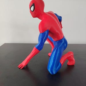 May include: A red, blue, and black Spider-Man figurine in a kneeling pose. The figure is primarily red with blue accents on the arms and legs. The mask has white eye lenses.