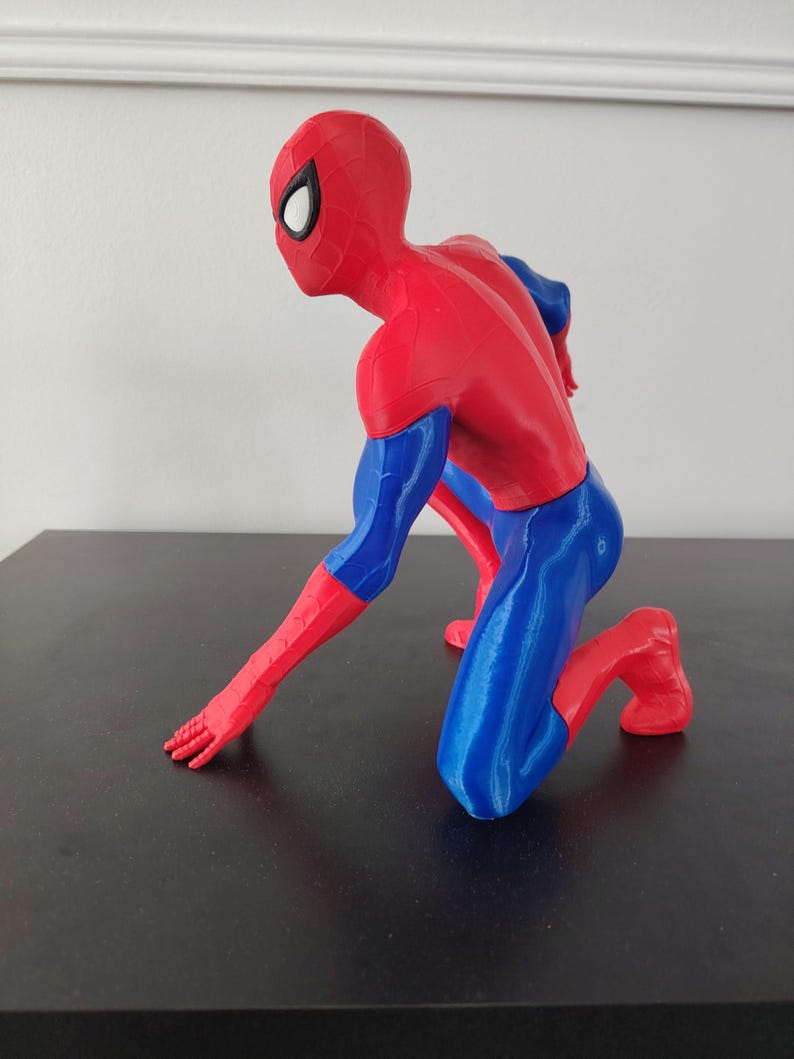 May include: A red, blue, and black Spider-Man figurine in a kneeling pose. The figure is primarily red with blue accents on the arms and legs. The mask has white eye lenses.