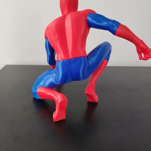 May include: A red and blue Spider-Man figurine in a crouching pose. The figure is viewed from the back, showcasing the iconic costume's colors and design. The figure is on a black surface.