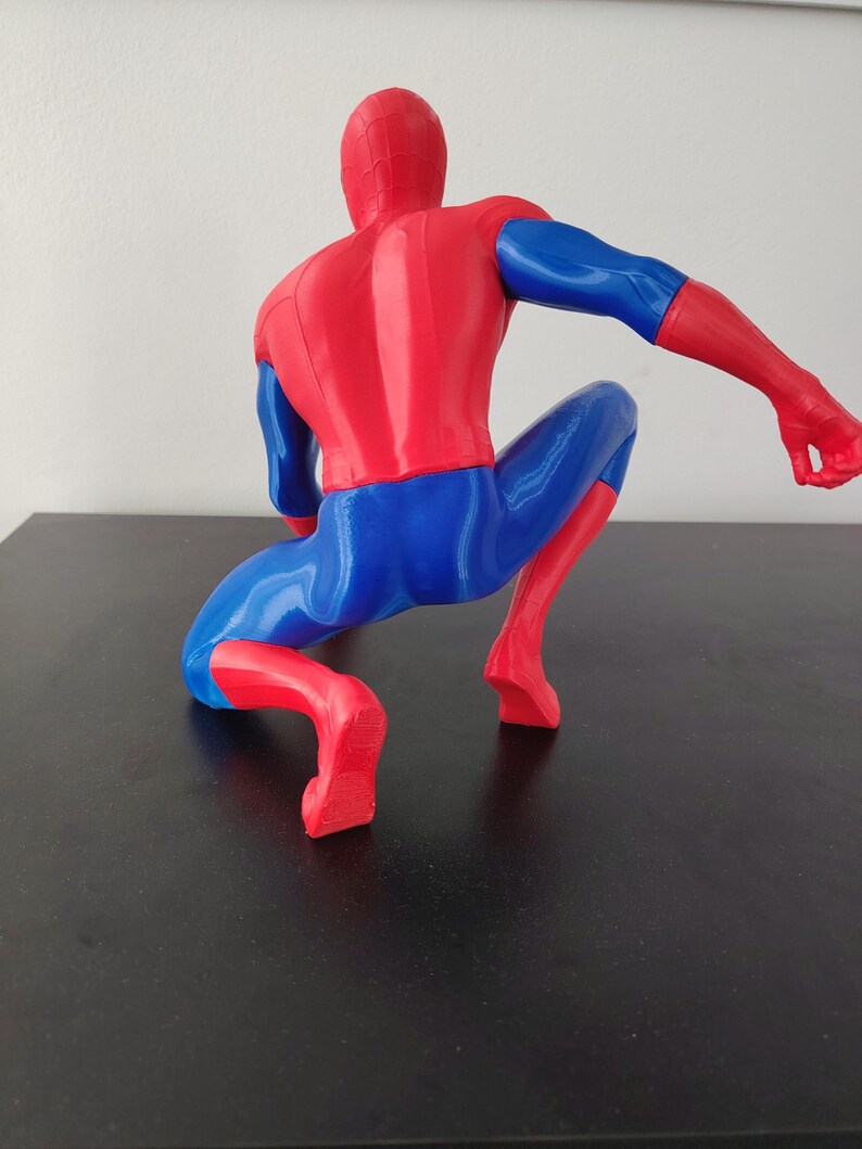 May include: A red and blue Spider-Man figurine in a crouching pose. The figure is viewed from the back, showcasing the iconic costume's colors and design. The figure is on a black surface.