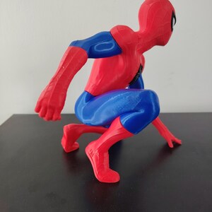 May include: A red, blue, and black Spider-Man figurine in a crouching pose. The figure has a red mask and suit with blue accents on the arms and legs. The figurine is on a black surface.