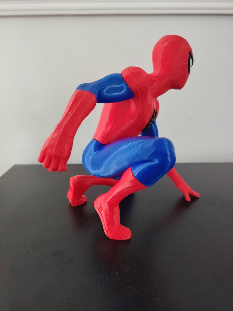 May include: A red, blue, and black Spider-Man figurine in a crouching pose. The figure has a red mask and suit with blue accents on the arms and legs. The figurine is on a black surface.