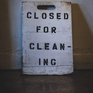 以下が含まれることがあります： 「CLOSED FOR CLEANING」の黒いステンシル文字が書かれた、風化した白い木製の看板。看板の上部には取っ手が切り抜かれており、壁に立てかけられています。