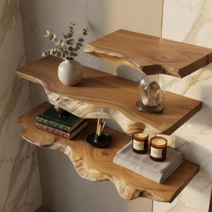 Op de afbeelding: Drie houten zwevende planken met natuurlijke randen zijn aan een muur bevestigd. De planken zijn versierd met een vaas, boeken, kaarsen en een glazen stolp. De planken zijn gemaakt van natuurlijk hout en hebben een warme, bruine tint.