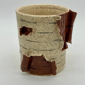 May include: A handmade ceramic mug with a speckled beige exterior and dark brown accents. The mug features a unique design with sections of the exterior appearing to peel away, revealing a darker brown interior. Black horizontal lines add detail.