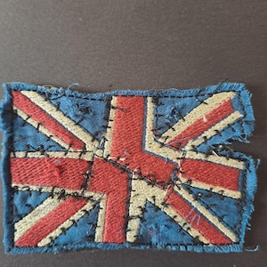 May include: A rectangular fabric patch featuring the Union Jack flag design in red, white, and blue. The patch has a frayed, raw edge and is constructed with visible, dark, irregular hand-stitched embroidery detailing across the surface.