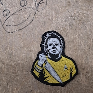 May include: An embroidered patch featuring a white-masked figure with messy hair wearing a yellow long-sleeved shirt with a delta-shaped emblem on the chest. The figure holds a large silver knife in one hand, with black outlines defining the design.