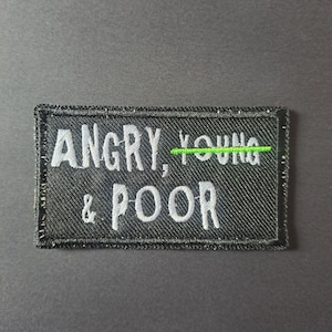 May include: A rectangular black embroidered patch featuring the white stitched text ANGRY, YOUNG & POOR. The word YOUNG has a horizontal bright green line stitched through the centre, crossing out the text with a single straight thread line.