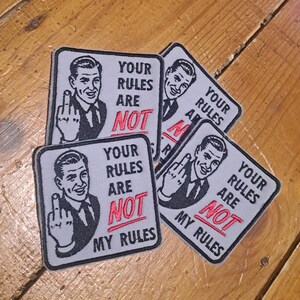 May include: Four square patches with a retro illustration of a man giving the middle finger. Each patch has the text "YOUR RULES ARE NOT MY RULES" in black and red on a grey background.