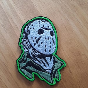 May include: An embroidered patch featuring a light blue hockey mask with black eye holes and ventilation dots. The mask is positioned above a dark green and black collar, with the entire design outlined in a vibrant neon green thread border.