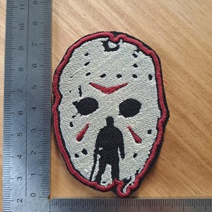 May include: An embroidered patch shaped like a hockey mask in off-white with black eye holes, red accents, and a black silhouette of a figure standing at the bottom. A metal ruler is visible along the left and bottom edges for scale.