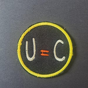 May include: A circular embroidered patch featuring a black background with a bright yellow border. The centre displays the white letters U and C separated by an orange equals sign, all rendered in a simple, hand-drawn style font.
