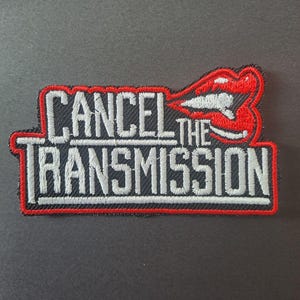 May include: An embroidered patch featuring the text CANCEL THE TRANSMISSION in white block letters with a black background and a red border. To the right of the text is an embroidered graphic of a pair of red lips with white teeth.