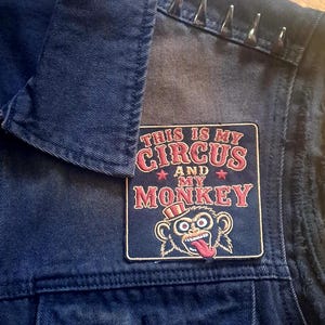 May include: A dark blue denim waistcoat featuring a rectangular patch with the red text "THIS IS MY CIRCUS AND MY MONKEY" above a graphic of a monkey wearing a top hat. The shoulder area is adorned with a row of silver-coloured metal cone studs.