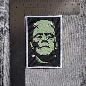 May include: A rectangular patch with a green Frankenstein monster design on a black background, framed by a white border. The patch measures approximately 7.6 cm by 6.4 cm.