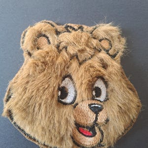 May include: A brown faux fur bear head patch featuring embroidered details. The bear has white and black eyes, a tan muzzle, a black nose, and a small open mouth with a red tongue. The edges are finished with black stitching in a rounded shape.