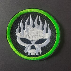 May include: A circular embroidered patch featuring a stylised white skull with flame-like details on a black background, enclosed by a vibrant green border. The design is stitched with dense thread, creating a textured appearance on the round fabric base.
