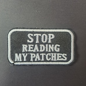 May include: A rectangular embroidered patch with a black background and a white border. The patch features the white embroidered text STOP READING MY PATCHES in a bold, sans-serif font, arranged in three lines centred on the fabric surface.