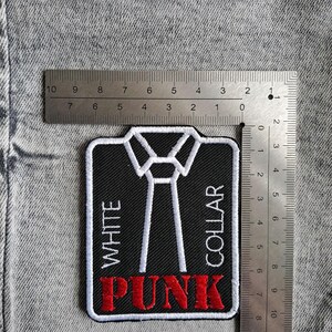 May include: An embroidered patch with a black background, white tie design, and the words "WHITE COLLAR PUNK" in white and red. The patch measures approximately 7.6 cm wide and 7.6 cm tall. The patch is on a grey denim background.