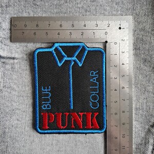 May include: Embroidered patch featuring a black background and a blue collar design. The words "BLUE COLLAR PUNK" are embroidered in blue and red. The patch measures approximately 7.6 cm by 7.6 cm.