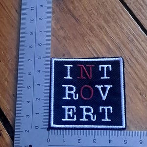May include: A square black embroidered patch with a white border featuring the text INTROVERT in white and red stitching. The patch is positioned against an L-shaped metal ruler, showing it measures approximately 5 cm by 5 cm in size.