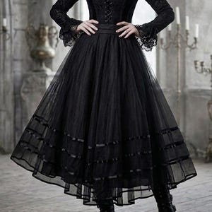Könnte beinhalten: Ein schwarzes Langarmkleid mit hohem Spitzenkragen, einem Korsett-Oberteil mit Schnürung vorne und einem voluminösen Tüllrock mit horizontalen Bandverzierungen. Das Kleid wird mit schwarzen Plateaustiefeln und einem Kopfschmuck mit Federn kombiniert.
