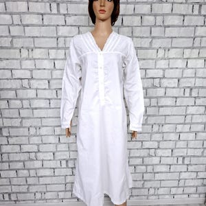 May include: A white long-sleeve nightgown with a V-neckline and button-down front. The nightgown features decorative trim along the neckline and a knee-length hem. The garment is displayed on a mannequin against a white brick wall.