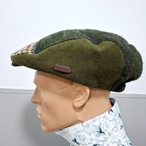 May include: A green tweed newsboy cap with a brown leather tag. The cap features a patchwork design with a solid green section and a patterned section in shades of green, brown, and beige. The cap is displayed on a mannequin head.