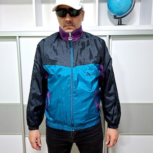 May include: A vintage Puma track jacket in black, teal, and purple. The jacket has a zip-up front, a purple collar, and the Puma logo on the chest. The jacket is worn with a white cap and sunglasses.