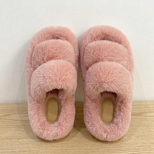 May include: A pair of soft, pink faux fur slippers. Each slipper has a thick, fluffy strap across the top and a light brown insole. The slippers are designed for indoor use and provide comfort and warmth.