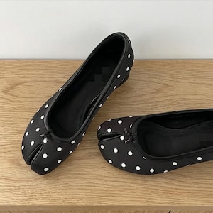 Op de afbeelding: Een paar zwarte ballerina's met witte stippen. De schoenen hebben een split-teenontwerp en een klein strikdetail aan de voorkant. Ze zijn gemaakt van een gladde stof en worden gepresenteerd op een houten oppervlak.