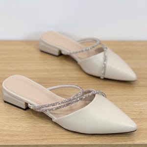 Könnte beinhalten: Ein Paar cremefarbene Mules mit spitzer Zehenpartie und kleinem Absatz. Die Schuhe haben einen Riemen über dem Fuß und einen zweiten Riemen, der mit glitzernden Strasssteinen verziert ist. Die Schuhe stehen auf einer hellen Holzoberfläche.
