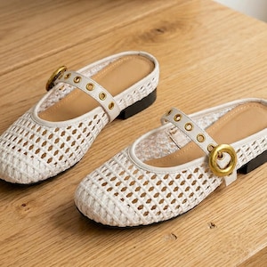 Puede incluir: Un par de zapatos blancos de cuero tejido con correa y hebilla dorada. Los zapatos tienen punta cuadrada y un pequeño tacón negro. El interior es de color canela.