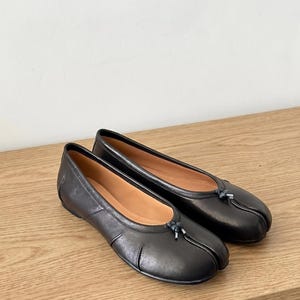May include: A pair of black leather ballet flats with a split-toe design. The shoes have a rounded toe and a small bow detail at the top. The interior is a light tan color.