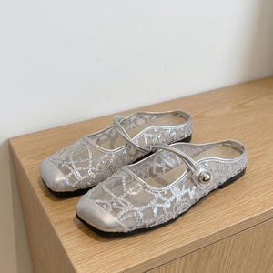 Puede incluir: Un par de bailarinas de malla plateada con puntera cuadrada y una correa en la parte superior. Los zapatos tienen un estampado floral decorativo y un pequeño botón plateado en la correa. Las suelas son negras.