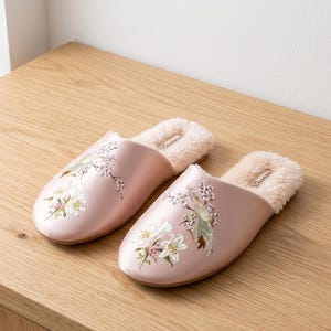 Pode incluir: Um par de chinelos de cetim rosa claro com bordado floral. Os chinelos têm um forro macio de cor creme e são exibidos em uma superfície de madeira. O bordado apresenta flores brancas e verdes.